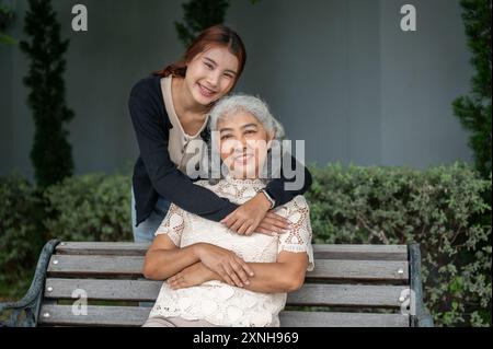 Eine hübsche asiatische Enkelin umschließt ihre Großmutter mit Liebe, während sie zusammen in einem Garten ausruht, beide lächeln in die Kamera. Familienverbundenheit, an Stockfoto