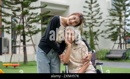 Eine hübsche asiatische Enkelin umarmt ihre Oma, die im Rollstuhl sitzt, während sie in einem Park läuft, Zeit miteinander verbringt und sich mit Liebe umschließt Stockfoto