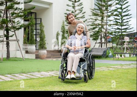 Eine reizende asiatische Enkelin und eine glückliche pensionierte Oma im Rollstuhl sind in ihrem wunderschönen Garten zu Hause, lächeln in die Kamera und nehmen Witz auf Stockfoto