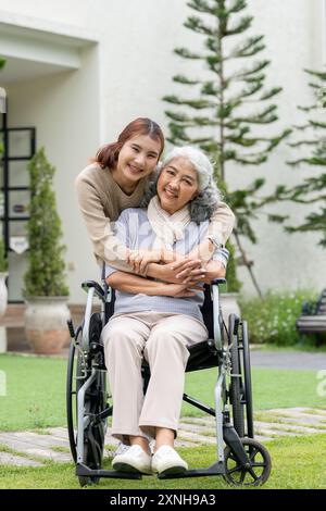 Eine reizende asiatische Enkelin und eine glückliche pensionierte Oma im Rollstuhl sind in ihrem wunderschönen Garten zu Hause, lächeln in die Kamera und nehmen Witz auf Stockfoto