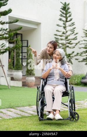 Eine reizende asiatische Enkelin und eine glückliche pensionierte Oma im Rollstuhl sind in ihrem wunderschönen Garten zu Hause, schauen zusammen die Aussicht und umarmen sie Stockfoto