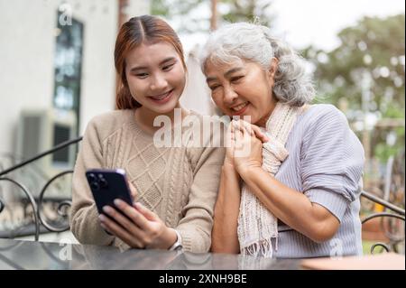 Eine hübsche asiatische Enkelin unterrichtet ihre Großmutter mit einem modernen Smartphone oder sieht sich gemeinsam Videos auf einem Smartphone an, während sie am Tisch sitzt Stockfoto