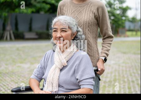 Eine lächelnde, reife asiatische Großmutter im Rollstuhl verbringt Zeit mit ihrer Tochter oder ihrem Betreuer in einem Park. Betreuer, Behinderte, Gesundheitsfürsorge Stockfoto