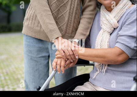 Ein Nahbild einer Tochter, die die Hand ihrer Mutter hält, während sie im Rollstuhl sitzt und ihre gemeinsame Zeit im Park genießt. Liebe, Bindung, Vertrauen, enco Stockfoto