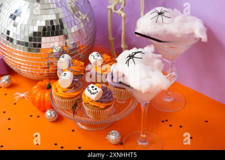 Halloween-Party-Komposition mit Cupcakes, Disco-Ball, Getränken und Dekorationen auf orangenem Tisch vor lila Hintergrund Stockfoto