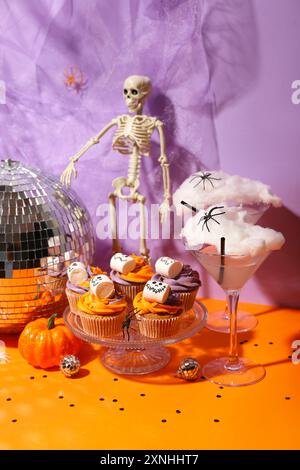 Halloween-Party-Komposition mit Cupcakes, Disco-Ball, Getränken und Dekorationen auf orangenem Tisch vor lila Hintergrund Stockfoto