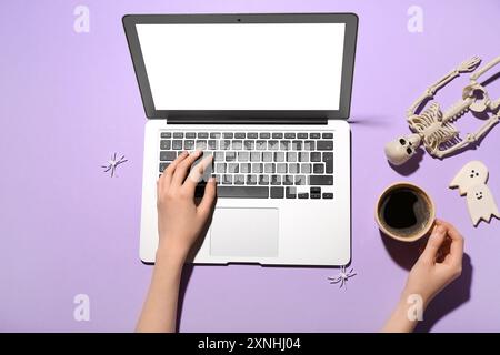 Frauenhände mit Laptop, Tasse Kaffee und Halloween-Deko auf fliedernem Hintergrund Stockfoto