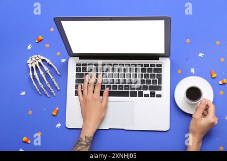 Weibliche Hände mit Laptop, Tasse Kaffee und Halloween-Deko auf blauem Hintergrund Stockfoto
