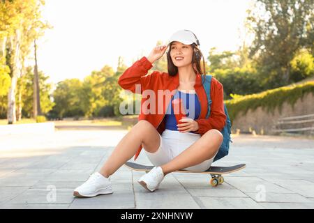 Junge asiatische Frau in Kopfhörern mit Sprühdose, die draußen auf Skateboard sitzt Stockfoto