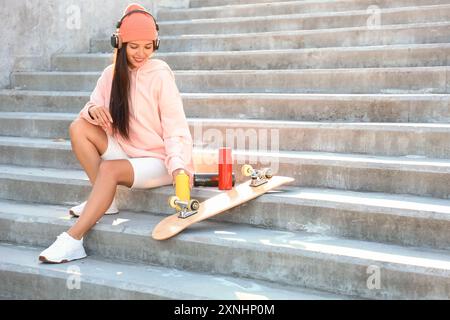 Junge asiatische Frau in Kopfhörern mit Sprühdose und Skateboard, die draußen auf Treppen sitzt Stockfoto