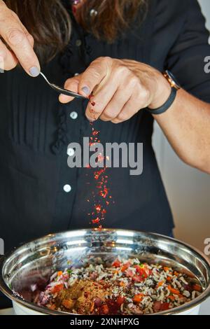 Nahaufnahme einer Person, die gemahlene Gewürze über einen bunten Salat in einer Edelstahlschüssel in der Küche streut. Stockfoto