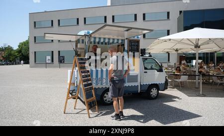 Ein Mann steht vor einem Imbisswagen und schaut sich die Speisekarte an. Der Lkw steht vor einem Gebäude mit Markisen und Sonnenschirmen. Es gibt mehrere p Stockfoto