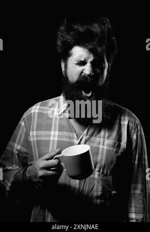 Mans hält eine Tasse Kaffee und Gähnen auf Schwarz. Morgentee. Gähngesicht. Mann mit Teetasse. Hipster-Mann gähnt mit einer Tasse Kaffee. Bärtiger Mann Stockfoto