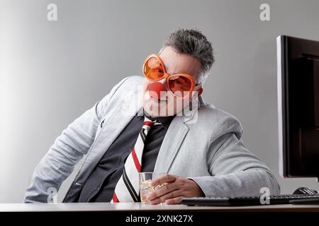 Geschäftsmann sitzt an seinem Schreibtisch und trinkt Alkohol, während er eine Clownnnase und eine Brille trägt, und versucht, glückliche Probleme des Alkoholismus in der Arbeitsplatte zu erscheinen Stockfoto