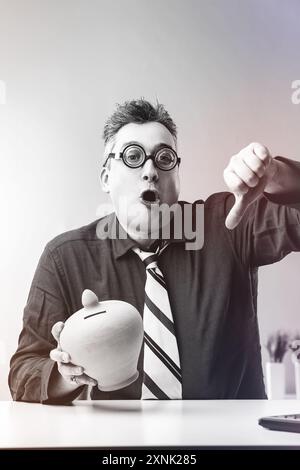 Ein Geschäftsmann hält eine Sparkasse und drückt mit schockiertem Ausdruck die Daumen nach unten, was auf finanzielle Probleme hindeutet Stockfoto