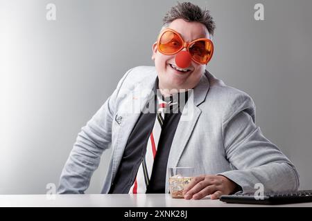 Geschäftsmann mit einer Clownnnase und lustiger Brille sitzt an einem Schreibtisch und trinkt Alkoholprobleme von Alkoholismus am Arbeitsplatz Stockfoto