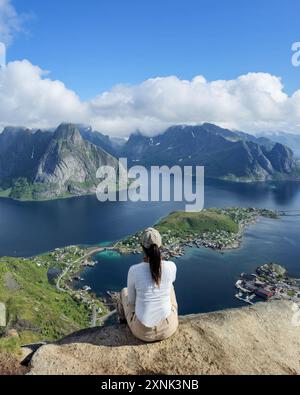 Ein Reisender sitzt auf einem felsigen Felsvorsprung und blickt unter hellem Himmel über das atemberaubende blaue Wasser und die zerklüfteten Berge der Lofoten. Reinebringen Lofoten Norwegen Stockfoto
