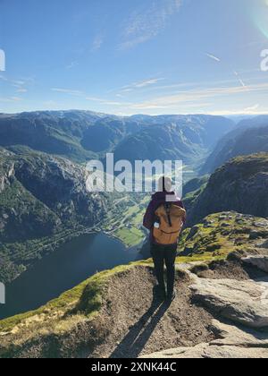 Ein einsamer Wanderer steht auf dem felsigen Felsvorsprung und blickt auf den atemberaubenden Fjord darunter, umgeben von majestätischen Bergen, die in Morgenlicht getaucht sind. Lysefjord Kjeragbolten Norwegen Stockfoto