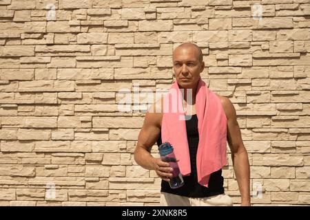 Porträt eines erwachsenen Athleten mit rosa Handtuch und Wasserflasche müde nach dem Training gegen beige Wand. Outdoor-Workout im Sommer. Kopierbereich. Stockfoto