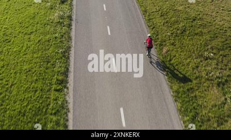 Rückansicht einer Rennradfahrerin, die auf einer leeren Asphaltstraße mit einem großen Schatten von Körper und Fahrrad fährt, Luftaufnahme einer Drohne. Stockfoto