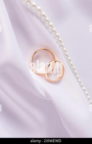 Schicker vertikaler Hochzeitshintergrund für Einladung Design, Karten, Cover. Ein Paar goldene Eheringe auf weißem Satin mit weichen Falten. Minimalismus Stockfoto