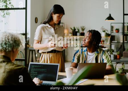 Junger Schwarzer Mann und asiatische Frau, die legere Outfits tragen und sich unterhalten, während der Programmierer im modernen Büro auf einem Laptop codiert Stockfoto