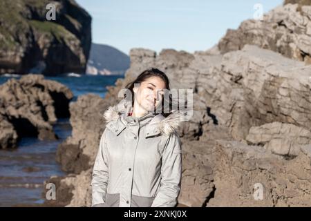 Eine Frau steht an einem felsigen Strand mit ihren Haaren im Wind. Trägt eine graue Jacke und lächelt Stockfoto
