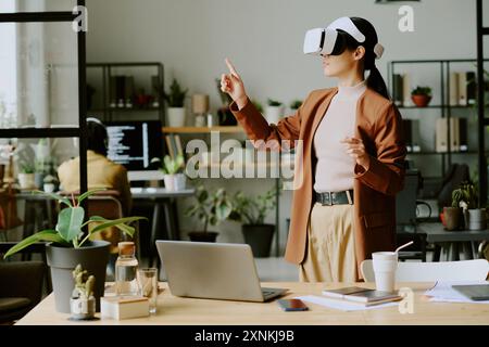 Junge asiatische Entwicklerin mit VR-Headset, die virtuelle Anwendung bei der Arbeit in führenden IT-Unternehmensbüros mit biophilem Design testen Stockfoto