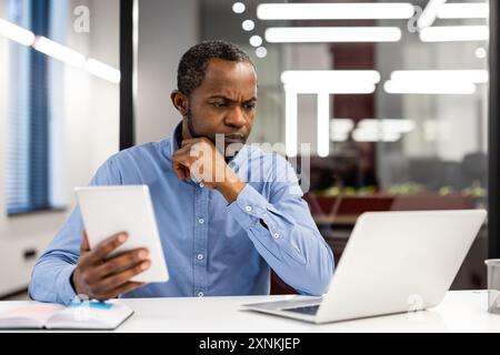 Ausgereifter afroamerikanischer Büromitarbeiter am Arbeitsplatz, der Daten auf Tablets und Laptops analysiert. Geschäftsmann konzentrierte sich auf die Arbeit in modernen Büros und zeigte Konzentration und Produktivität. Stockfoto