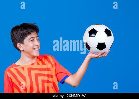 Das Kind hält den Fußball mit einer Hand und schaut ihn an. Isoliert auf blauem Hintergrund. Sport. Stockfoto