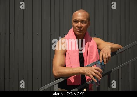 Porträt eines erwachsenen Athleten mit rosa Handtuch und Wasserflasche müde nach dem Training gegen dunkle Wände. Outdoor-Workout im Sommer. Kopierbereich. Stockfoto