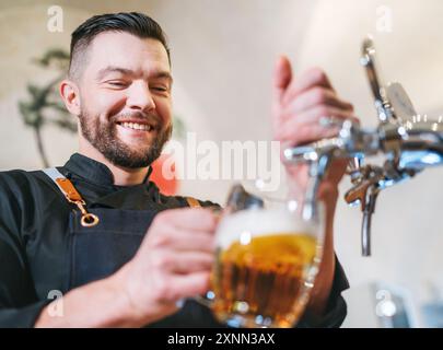 Ein lächelnder, stilvoller Bärenbarmann in schwarzer Uniform mit einer Schürze, der frisches Lagerbier in einen Pilsner-Glas-Becher an der Bar zapft. Erfolgreiche Mitarbeiter, Stockfoto