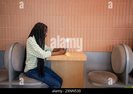 Afroamerikanerin mit geflochtenem Haar und Brille sitzt im modernen Café, konzentriert auf ihr Smartphone und genießt einen Moment der Konnektivität und Entspannung. Stockfoto