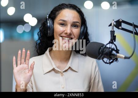 Lächelnde Frau mit Kopfhörern, die die Hand winkt, während sie Podcasts im Studio aufnimmt. Weibliche Moderatorin sitzt am Mikrofon und blickt in die Kamera und genießt die Übertragung. Konzeptkommunikation, Medien, Podcasting. Stockfoto