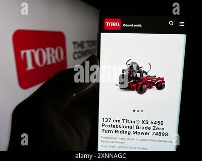 Person, die ein Mobiltelefon mit einer Webseite des US-Rasenmähergeschäfts The Toro Company vor dem Geschäftslogo hält. Konzentrieren Sie sich auf die Mitte des Telefondisplays. Stockfoto