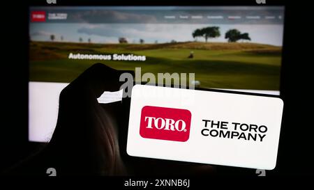 Person, die ein Mobiltelefon mit dem Logo des amerikanischen Rasenmäher-Unternehmens The Toro Company vor der Business-Webseite hält. Schwerpunkt auf dem Display des Telefons. Stockfoto