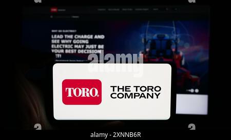 Person, die ein Mobiltelefon mit dem Logo des US-Rasenmähergeschäfts The Toro Company vor der Business-Website hält. Schwerpunkt auf dem Display des Telefons. Stockfoto