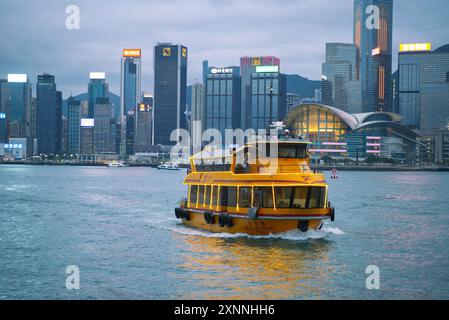 Gelbes touristisches Kreuzfahrtschiff, das in der Abenddämmerung im Victoria Harbour mit der Skyline dahinter segelt. Hongkong - 27. Mai 2024 Stockfoto