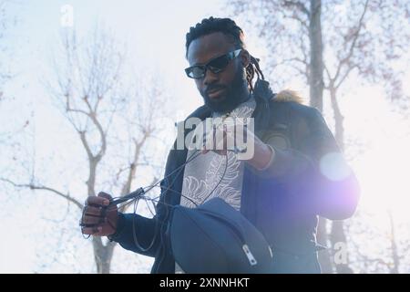 Junger afrikanischer Mann, der im Park steht und ein Sprungseil aus der Bauchtasche herausnimmt, Sonnenbrille und Kleidung für kaltes Wetter trägt, das Bild hat eine Atmosphäre mit Sonnenliedern Stockfoto