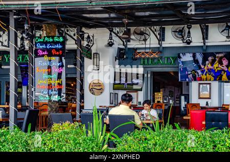 Eine Familie isst in einem Restaurant am Straits Quay in Penang, Malaysia. Ein farbenfrohes Schild in Schwarz und Neon zeigt einige der verfügbaren Menüoptionen an. Stockfoto