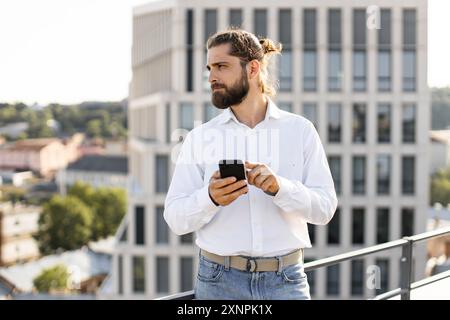 Mann in weißem Hemd mit Smartphone auf dem Dach mit Blick auf die Stadt Stockfoto