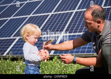 Der moderne Vater und sein Kind legen Geld in die Sparkasse. Das kleine Kind will Geld für neues Spielzeug sparen. Mann und sein Sohn, die Geld in die Sparkasse auf dem Hintergrund einer Solarbatterie stecken. Stockfoto