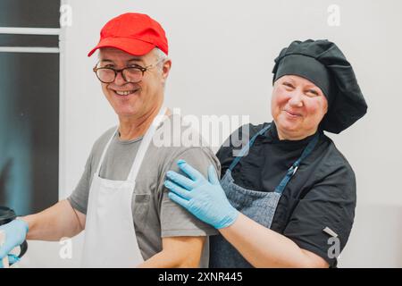 Ein fröhlicher älterer Mann in roter Mütze und Brille, trägt eine Schürze und steht neben einer lächelnden Frau mit Kochmütze und Schürze. Sie sind in einer Küche setti Stockfoto