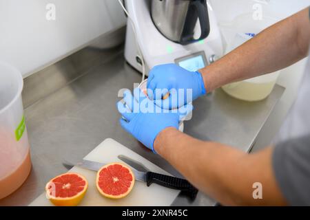 Eine Person, die blaue Handschuhe trägt, bereitet Grapefruit in einer Küche zu. Sie schneiden eine Grapefruit auf einem weißen Schneidebrett, mit einem Messer daneben Stockfoto