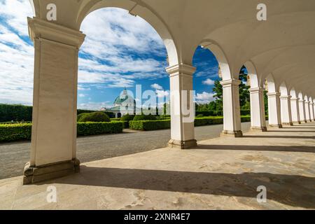 Blick durch die Arkaden der barocken Kolonnade mit Statuen alter Götter und Schatten an einem sonnigen Tag auf den dekorativen Blumengarten und das Gebäude Stockfoto