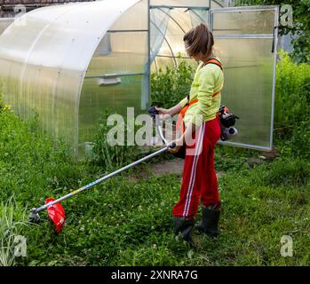 Eine junge Frau mit einem Benzintrimmer mäht an einem Sommerabend das Gras auf dem Rasen vor dem Gewächshaus in einem Landhaus Stockfoto