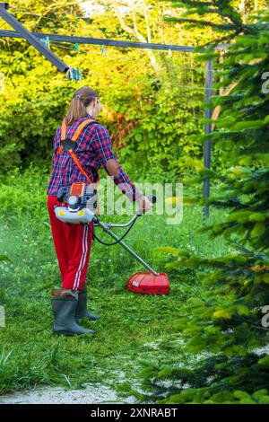 Eine junge Frau mit einem Benzintrimmer mäht an einem Sommerabend das Gras auf dem Rasen in einem Landhaus Stockfoto