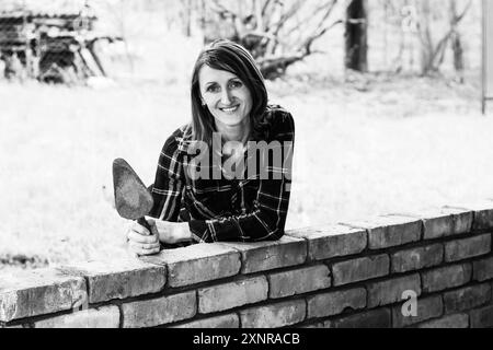 Eine junge Maurerin mit einer Kelle in den Händen lächelt und lehnt sich an eine Ziegelwand, ein Schwarzweißfoto Stockfoto