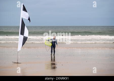 Eine reife Surferin, die ihr Surfbrett trägt und am Towan Beach in Newquay in Cornwall in Großbritannien aus dem Meer wandert. Stockfoto