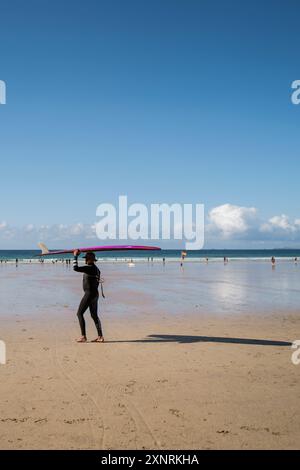 Ein Surfer mit Hut und balanciertem Surfbrett auf dem Kopf, der bei Ebbe am Towan Beach in Newquay in Cornwall in Großbritannien spaziert. Stockfoto